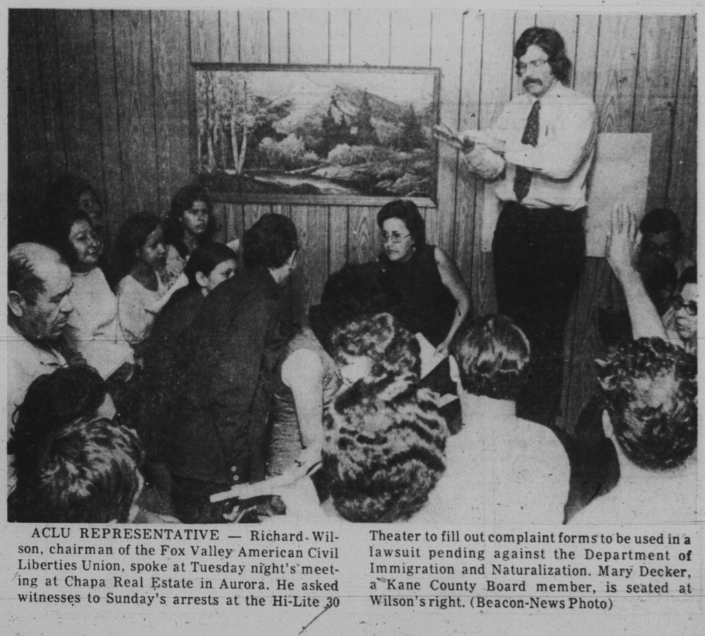Clipping of a newspaper article image. Photo is black and white and shows a number of folks in a room with wood paneling listening to a seated Mary Decker and a man standing next to her.